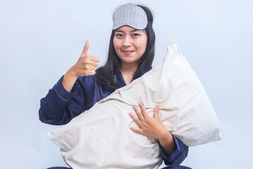 Young Asian woman in pajamas and sleep eye mask hold pillow and showing thumbs up over grey...
