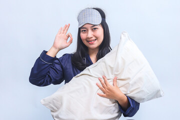 Young Asian woman in pajamas and sleep eye mask hold pillow and showing okay hand gesture on grey...