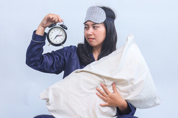 Young woman in pajamas and sleep eye mask hold alarm clock isolated on gray background. Night...
