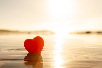 A resin heart rests on the wet beach sand, touched by the waves and illuminated by the golden light of sunrise. This image evokes feelings of romance and love