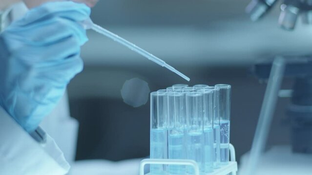 Young female microbiologist in disposable gloves and safety glasses adding liquids in test tubes with pipette while performing experiment in medical laboratory. Close-up view, tilt-up shot