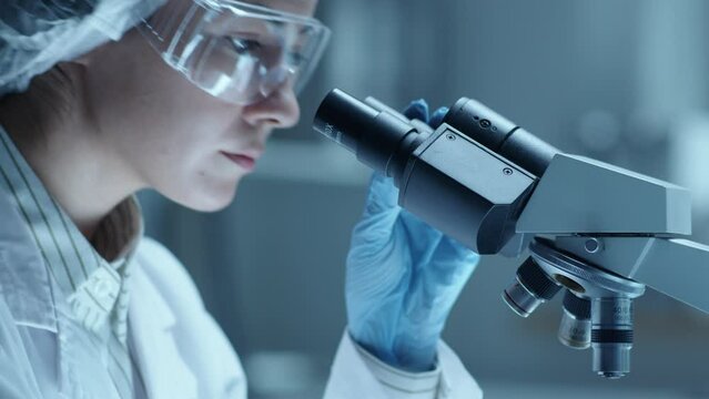 Female scientist in safety glasses, sterile hat and gloves placing glass slide with specimen under microscope and performing medical test in laboratory. Tilt-up shot, close-up view