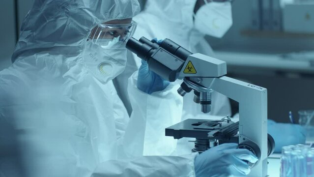 Female microbiologist wearing protective coverall suit, face mask, safety glasses and gloves performing microscopic examination of contagious blood sample in laboratory