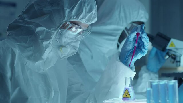 Female chemist wearing protective coverall suit, mask, glasses and gloves using pipette and dripping purple reagent into flask with transparent biohazard liquid during experiment in laboratory