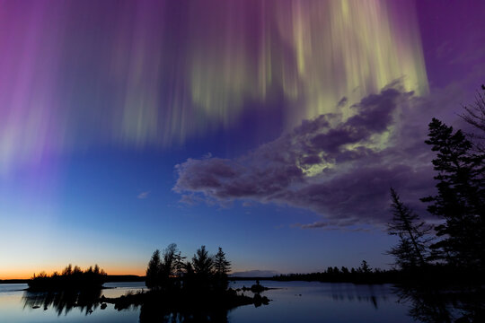 Northern lights erupt over a lake in Minnesota in a dark sky overhead shining rainbow of Aurora light as the sun sets on the horizon