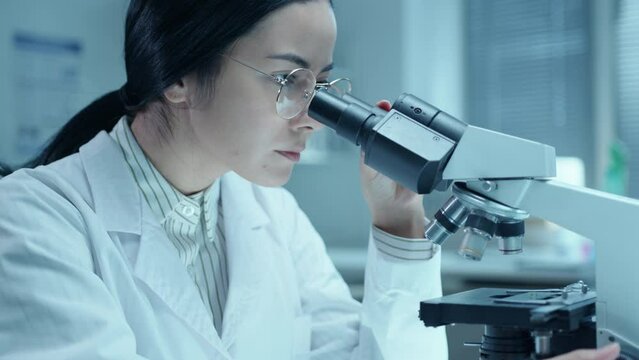 Young female doctor adjusting compound microscope and examining specimen in medical laboratory. Tilt-down shot