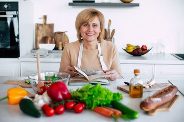 Close up portrait of beautiful smiling mature woman in apron is reading a book with recipes while cooking in the modern light kitchen with many vegetables on the table