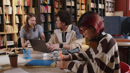 Young Asian female student with pink hair sitting at desk in college library, reading notes and marking text with highlighter pen while preparing for exams