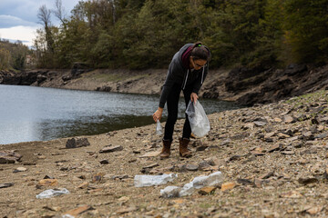Young female volunteer collecting plastic bottles from a lake shore or river bank, with forest in...