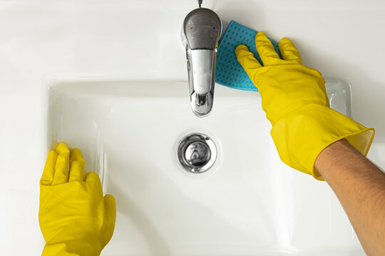 Hygiene concept aerial close up view of female hands wearing yellow protective rubber gloves cleaning a white modern ceramic sink around chrome tap or faucet and fittings using a blue sponge cloth