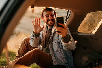 A man sits in the trunk of a car, waving and talking on a video call