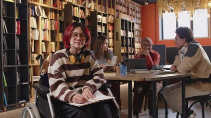 Portrait of gen Z female student in wheelchair taking notes in copybook and posing for camera with smile while doing homework in public library, diverse group mates sitting in background - Powered by Adobe