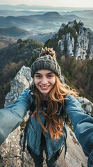 Naklejka premium A woman wearing a blue jacket and a knit hat is smiling and taking a selfie. Concept of adventure and excitement, as the woman is posing on a mountain top