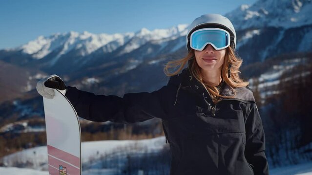 Woman snowboarder at snowy mountains. Happy woman snowboarder in costume and goggles with snowboard in hands at snowy mountains background. Having good time in holiday. Active extreme riding.