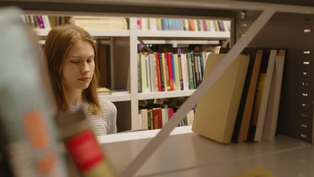 Young female student taking book from shelf and reading it in college library