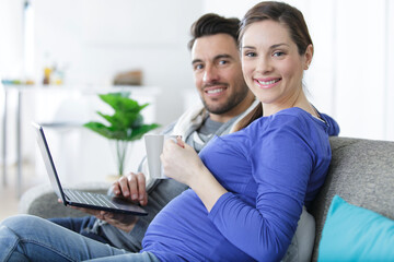 pregnant woman with husband using laptop while sitting at sofa