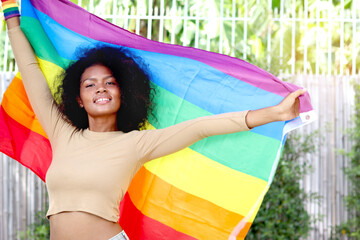 Happy smiling homosexual lesbian African woman with curly hair holding rainbow flag. Beautiful girl celebrating pride month outdoor. Cheerful LGBT and gay pride celebration movement concept.