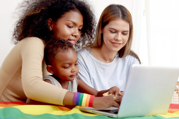Happy LGBT family, homosexual lesbian couple with rainbow flag wristband and adopted child use laptop computer together. Beautiful woman and African girlfriend search for pride celebration gifts.