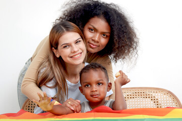 Happy homosexual lesbian couple with rainbow flag wristband and adopted child hug each other....