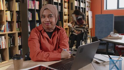 Portrait of positive Muslim girl in hijab sitting at desk with laptop and posing for camera with smile while studying in college library