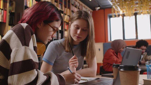 Two young gen Z female students sitting together at desk in college library, taking notes in workbook and talking while doing homework or getting ready for exam