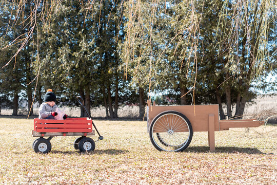 Toddler reading a newspaper in a red wagon under willow trees