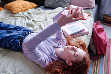 Modern teen girl wearing casual clothes lying on bed in her room taking selfies on smartphone, high...