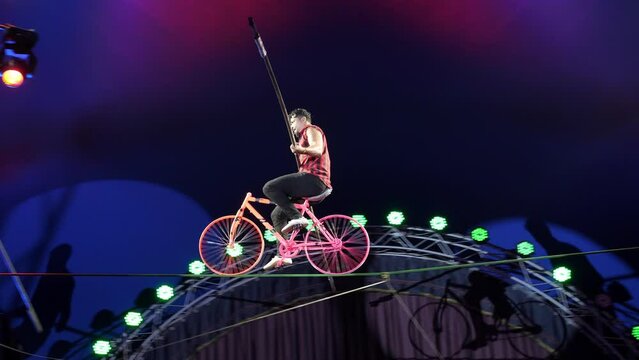An acrobat rides a bicycle on a tightrope under the dome of the circus. Performance of aerial gymnasts-tightrope walkers in a circus under the light of colorful spotlights. A circus show.