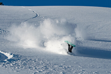 A snowboarder sticks upside down from the snow, feet up, having fallen in a huge cloud of snow against the backdrop of a lonely footprint on an untouched slope and the blue sky