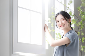 窓辺に立つ外の方を見上げる女性　朝日の中リラックスや早起きのイメージ　春夏の半袖　カメラ目線