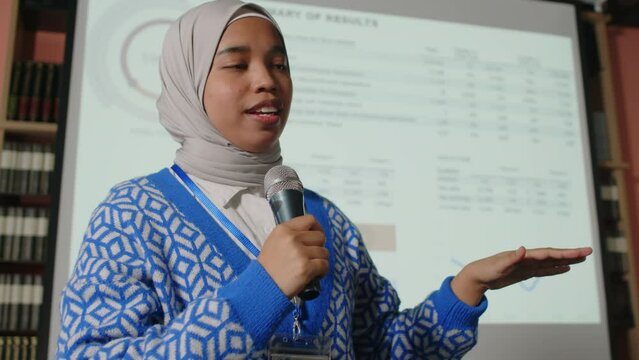 Young Muslim female speaker in Hijab pointing at presentation slide on projection screen and talking in microphone in front of audience during educational seminar in library