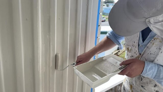 Middle-aged caucasian man in white painter's overalls uses a small paint roller to apply a neutral paint colour to old-fashioned fibre cement cladding in a sheltered porch area of a New Zealand home