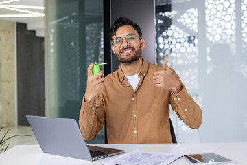 Man using sore throat spray and giving thumbs up while working on laptop in modern office