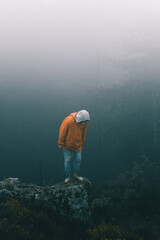Bulgaria - 05 22 2024: Man with orange jacket and blue jeans standing on the edge of a big rock looking down the cliff with dark and foggy and rainy background pine forest.