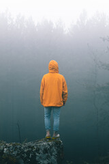 Bulgaria - 05 22 2024: Man with vibrant orange jacket and jeans standing on the edge of a rock looking at the bright light over the trees with a lot of fog.