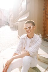 Fashionable young woman with a short haircut  in good mood posing sitting on the street stairs. Lifestyle, fashion, travel concept.