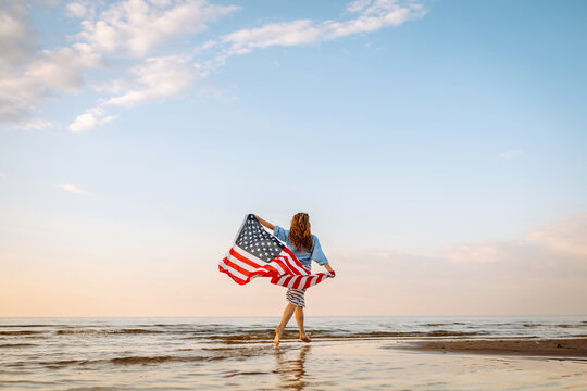 Young woman holding national American flag cover ocean beach holiday travel at summer romantic sunset. America independence day concept. 4th of July. - Powered by Adobe
