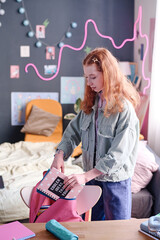 Red-haired girl standing in her room packing her backpack before going to school, vertical medium...