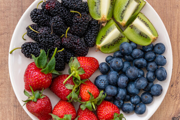 Close-up top view of blueberries, strawberries, blackberries and sliced kiwi fruit on a plate on the wooden surface