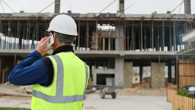 Building engineer talking on smartphone on construction site call with client, deadline, smart manager in helmet, back side view.