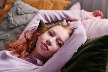 Medium closeup of red-haired teen girl relaxing on bed in her bedroom at home looking at camera