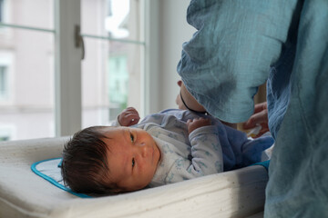 Mother changing her newborn baby's diaper on a changing table by a sunny window. The scene captures...