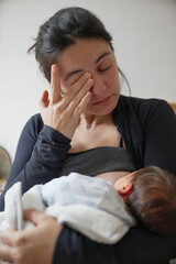 New mother wiping her eyes, holding her baby close while breastfeeding, illustrating the emotional...
