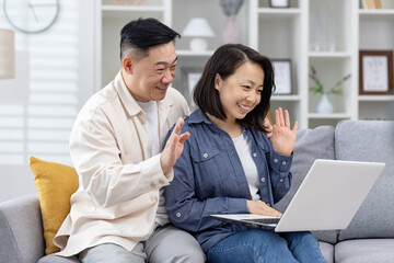 Cheerful couple enjoying a video call on their laptop while sitting on a couch at home, waving and smiling. - Powered by Adobe