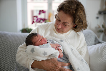 Mother holding newborn baby over her shoulder, standing near a bright window. The image captures a...