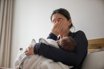 Mother breastfeeding her baby with a weary expression, covering her face partially, highlighting...