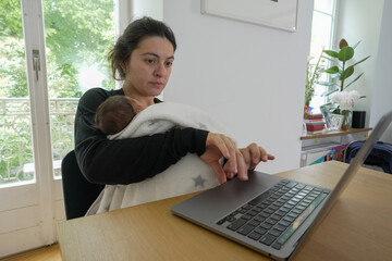 Mother multitasking while holding newborn baby, working on a laptop at home. She is focused on the...