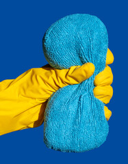 Hand in a Yellow Rubber Glove Squeezing a Dry Sponge. Hand in a Yellow Latex Glove with Blue Sponge on a Blue Background. 