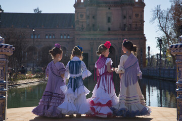 Four girls dancing flamenco, with their backs turned and talking to each other, in typical flamenco costumes next to a lake in a beautiful square in Seville. Concept dance, flamenco, typical Spanish.