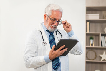 Mature senior male doctor in medical uniform using digital tablet in clinic. Therapist professional...